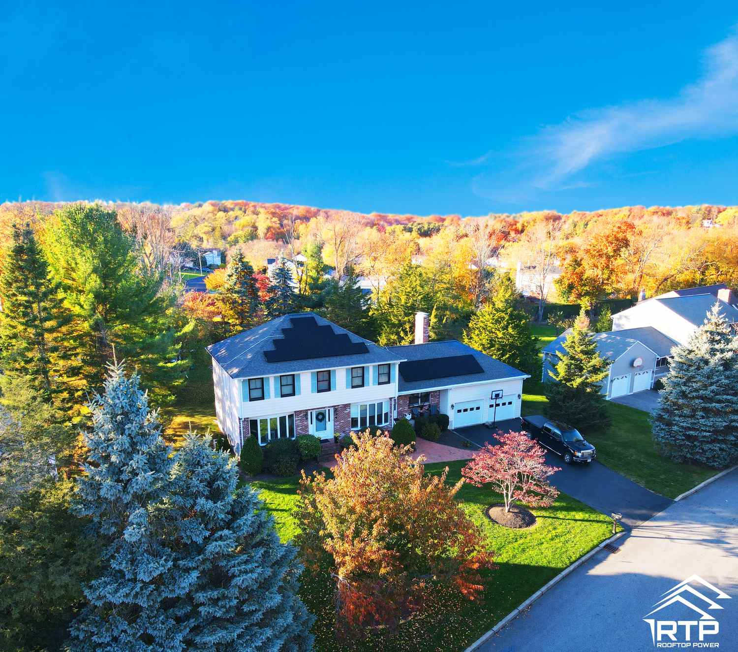 Aerial view of a white colonial farmhouse with black shutters, solar panels on two roof faces, surrounded by fall foliage, RTP logo visible in corner.