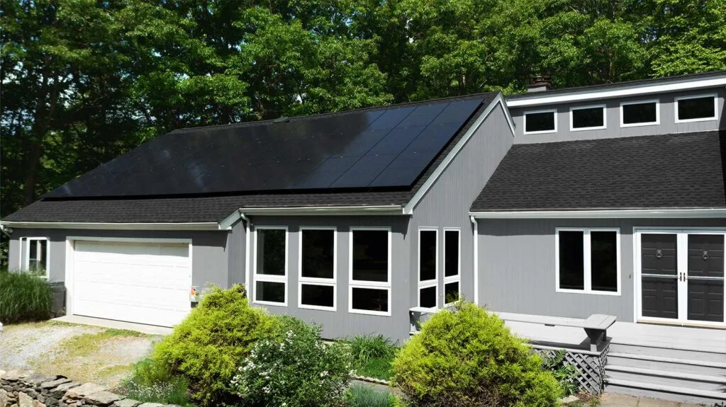 Ground-level view of a gray shingle-style contemporary home with a black solar panel array covering the front-facing roof slope, evergreen trees behind.