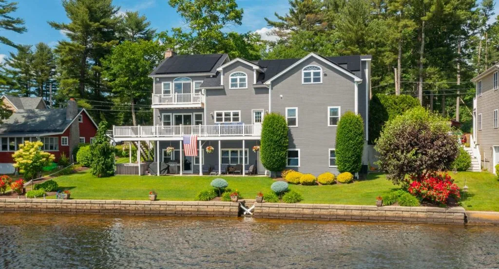 A gray three-story colonial home with solar panels on two roof faces, seen from the water, with a private dock and an American flag hanging from the deck.