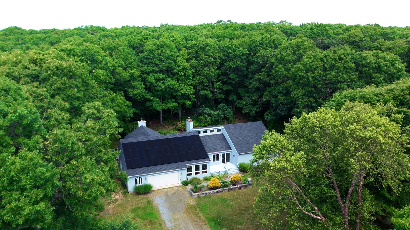Aerial view of a light blue cape-style home surrounded by dense New England forest, with black solar panels covering the front roof slope.