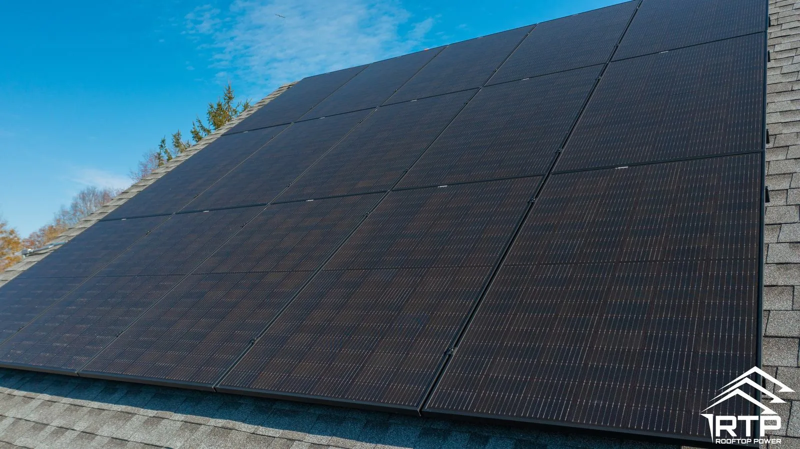 Close-up angle of an all-black solar panel array on an asphalt shingle roof, with fall foliage and blue sky in the background, RTP logo visible in corner.